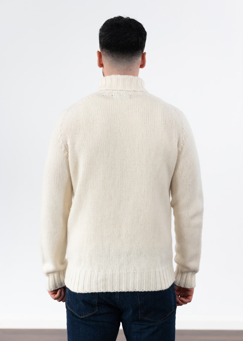 A man viewed from behind wearing a cream polo neck jumper and dark blue jeans, standing against a plain white studio background.