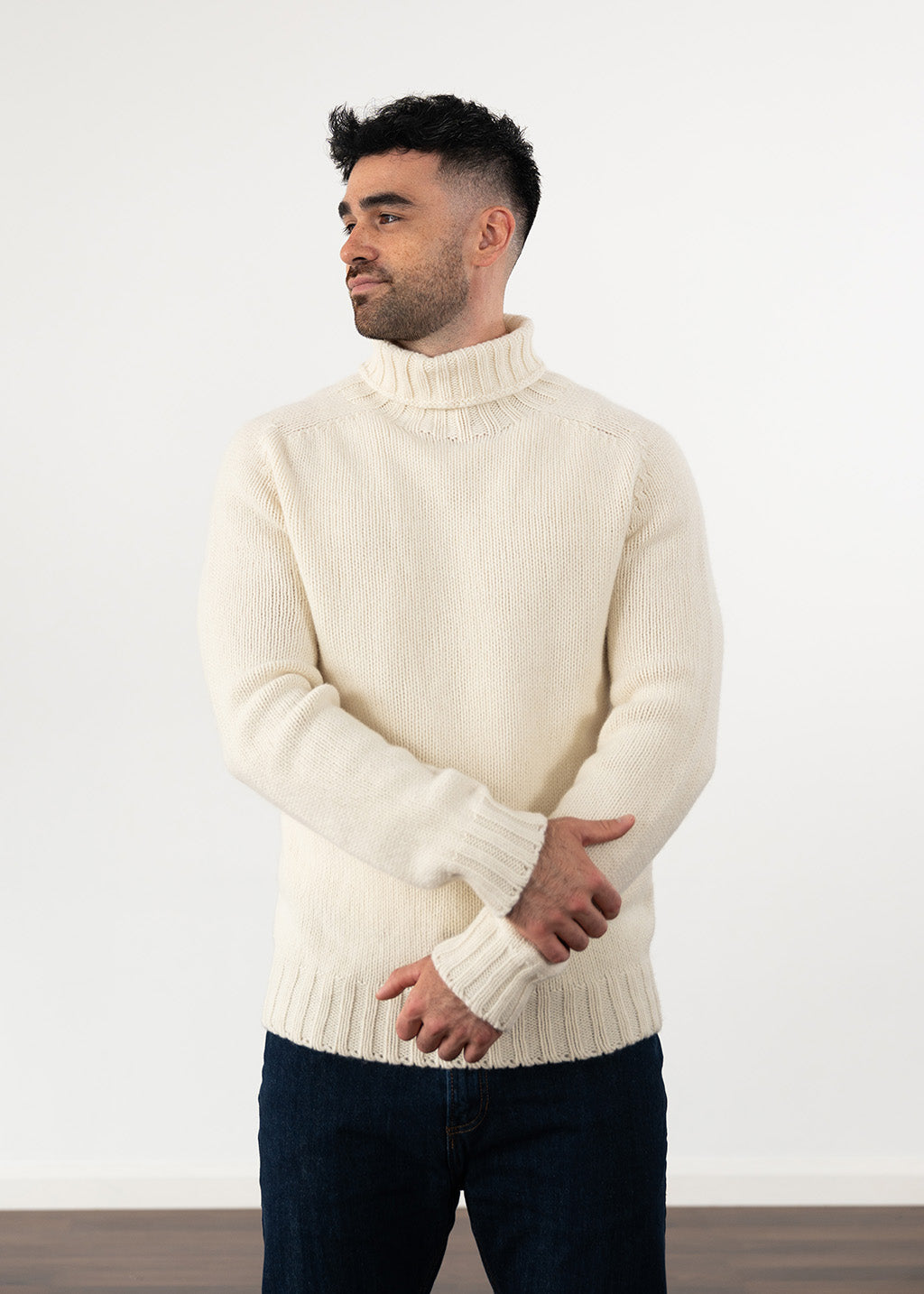 A man wearing a cream-coloured wool polo neck jumper and dark blue jeans, standing against a plain white wall.