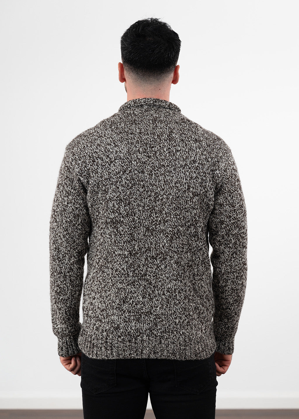 A man viewed from the back wearing a textured, marled knit sweater in black and white, paired with black jeans, against a plain white studio background.