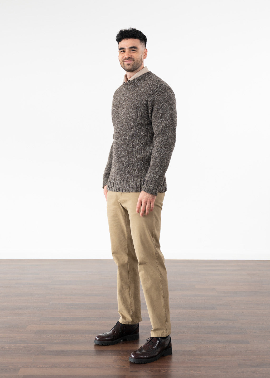 A man wearing a textured brown knit jumper, beige trousers, and dark brown leather shoes, standing in profile against a white studio background with a wood floor.