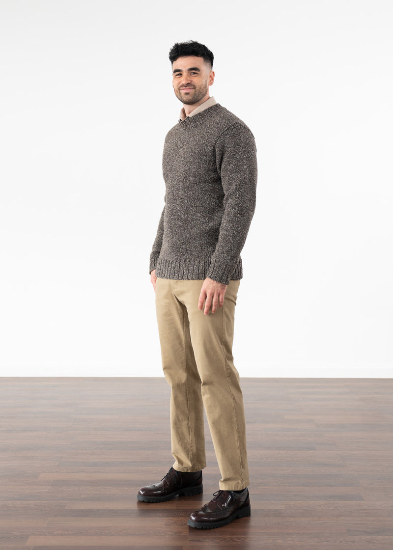 A man wearing a textured brown knit jumper, beige trousers, and dark brown leather shoes, standing in profile against a white studio background with a wood floor.