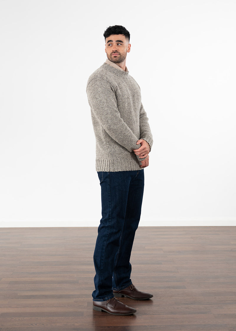 A man wearing a light grey-brown knit jumper, dark blue jeans, and brown leather shoes, standing in profile against a white studio background with a wooden floor.