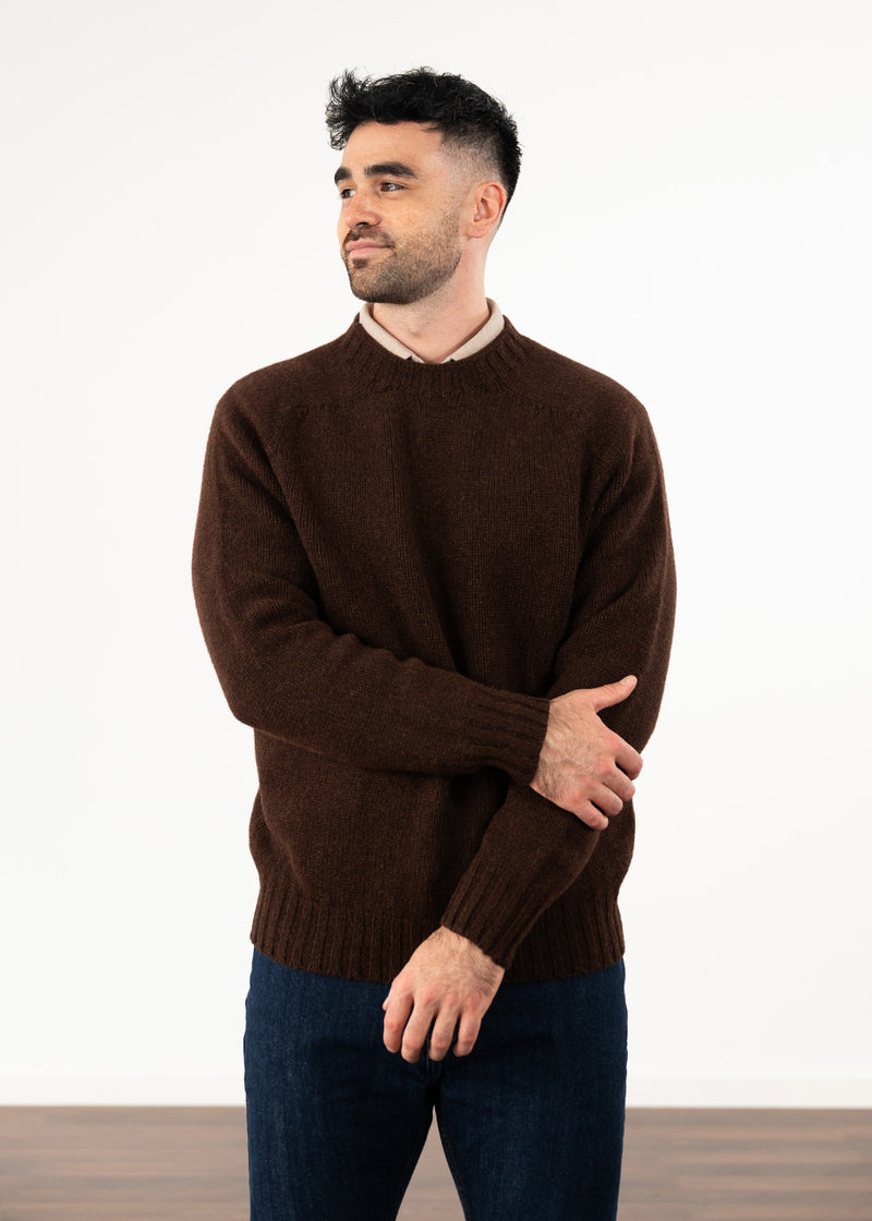 A man wearing a dark brown crew neck knit jumper over a light beige collared shirt and dark blue jeans, standing against a plain white wall.