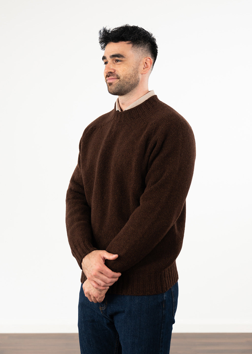 A man wearing a dark brown crew-neck knit sweater and dark blue jeans, standing in profile against a plain white studio background.