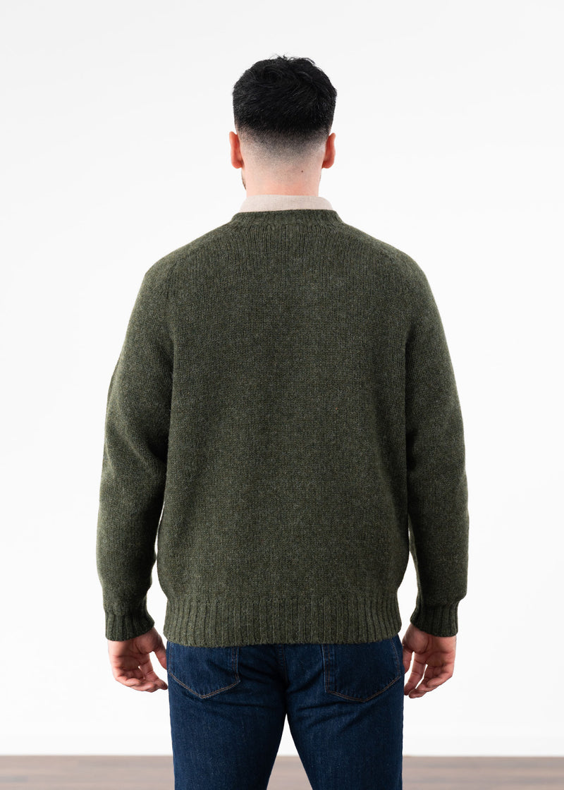 A man viewed from behind wearing an olive green knit sweater and dark blue jeans against a plain white studio background.