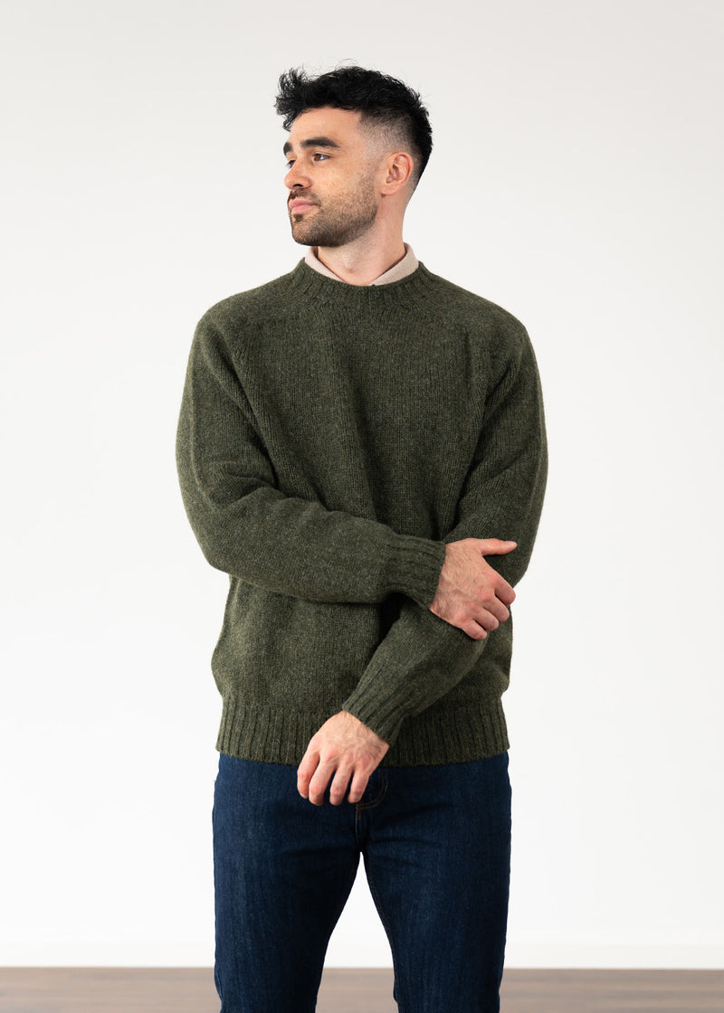 A man wearing a dark olive green knit crew neck jumper and dark blue jeans, standing against a plain white studio background.