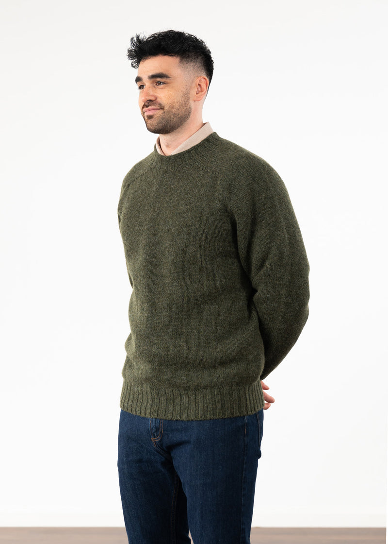 A man wearing an olive green knit crewneck sweater and dark blue jeans, standing against a plain white studio background.