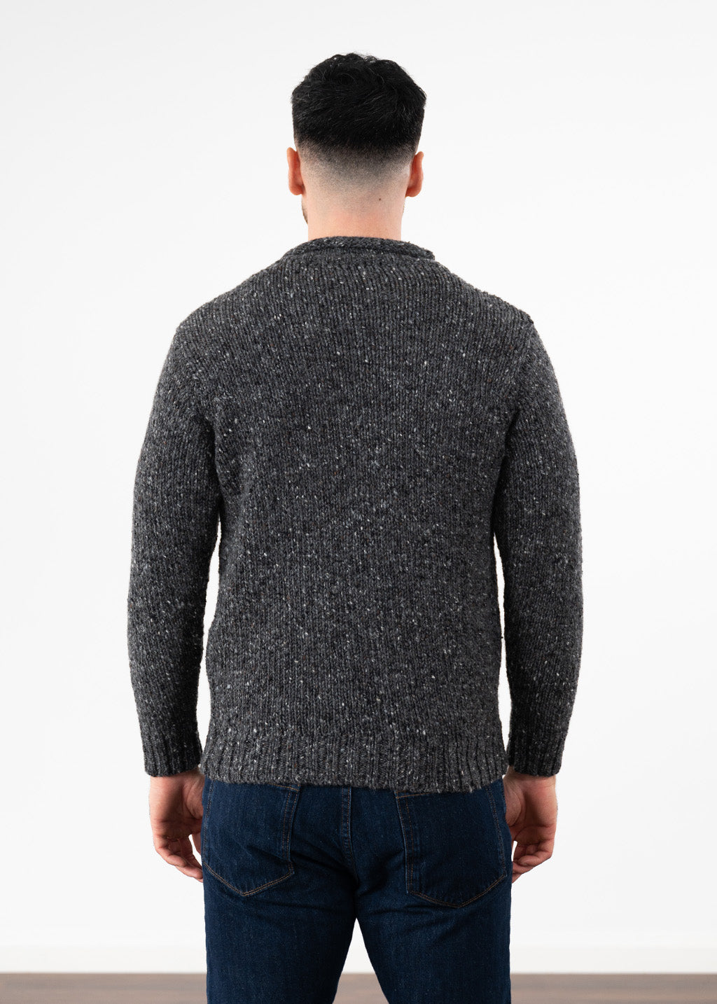 A man viewed from behind wearing a dark gray and black flecked knit jumper and dark blue jeans, standing against a plain white wall.