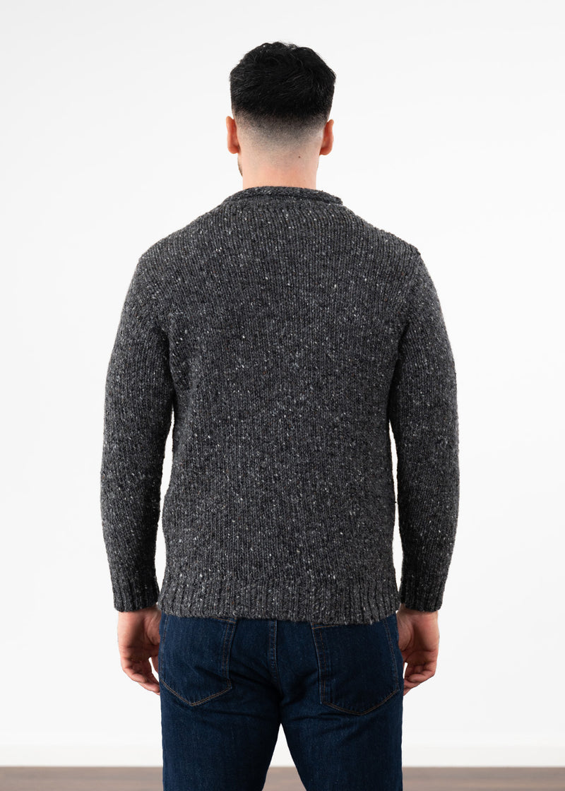 A man viewed from behind wearing a dark gray and black flecked knit jumper and dark blue jeans, standing against a plain white wall.
