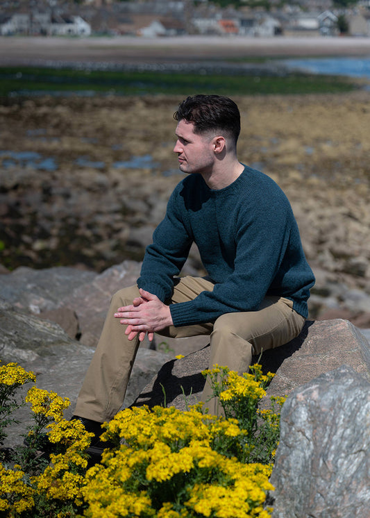 Man sitting on rocks by the shore wearing a dark teal Shetland jumper and beige chinos