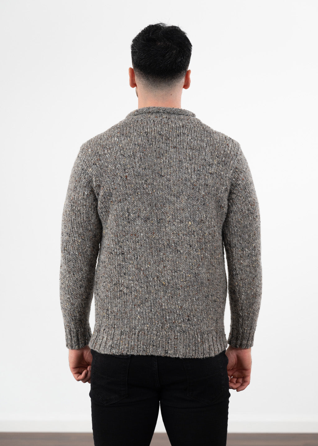 A man viewed from behind wearing a textured grey donegal wool knit jumper and black jeans, standing against a plain white wall.