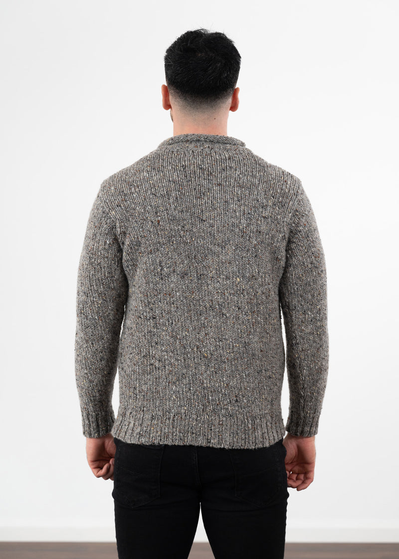 A man viewed from behind wearing a textured grey donegal wool knit jumper and black jeans, standing against a plain white wall.