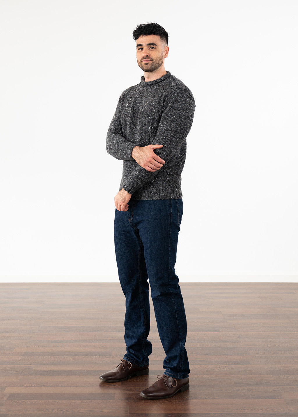 A man wearing a dark gray heathered knit jumper, dark blue jeans, and brown leather shoes, standing in a studio with a white wall and wooden floor.