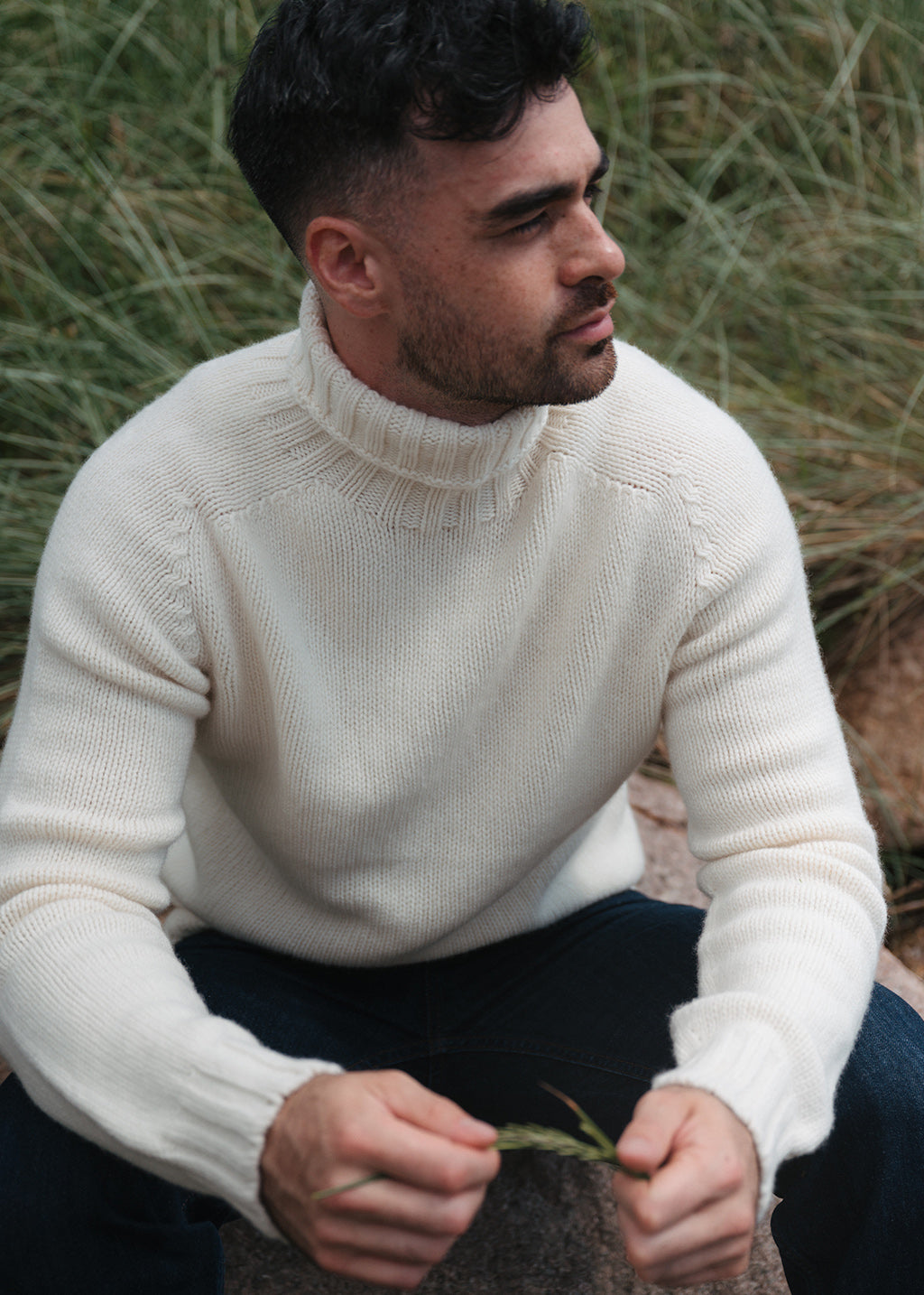 A man wearing a cream undyed wool polo neck jumper, sitting amongst  tall coastal marram grass.