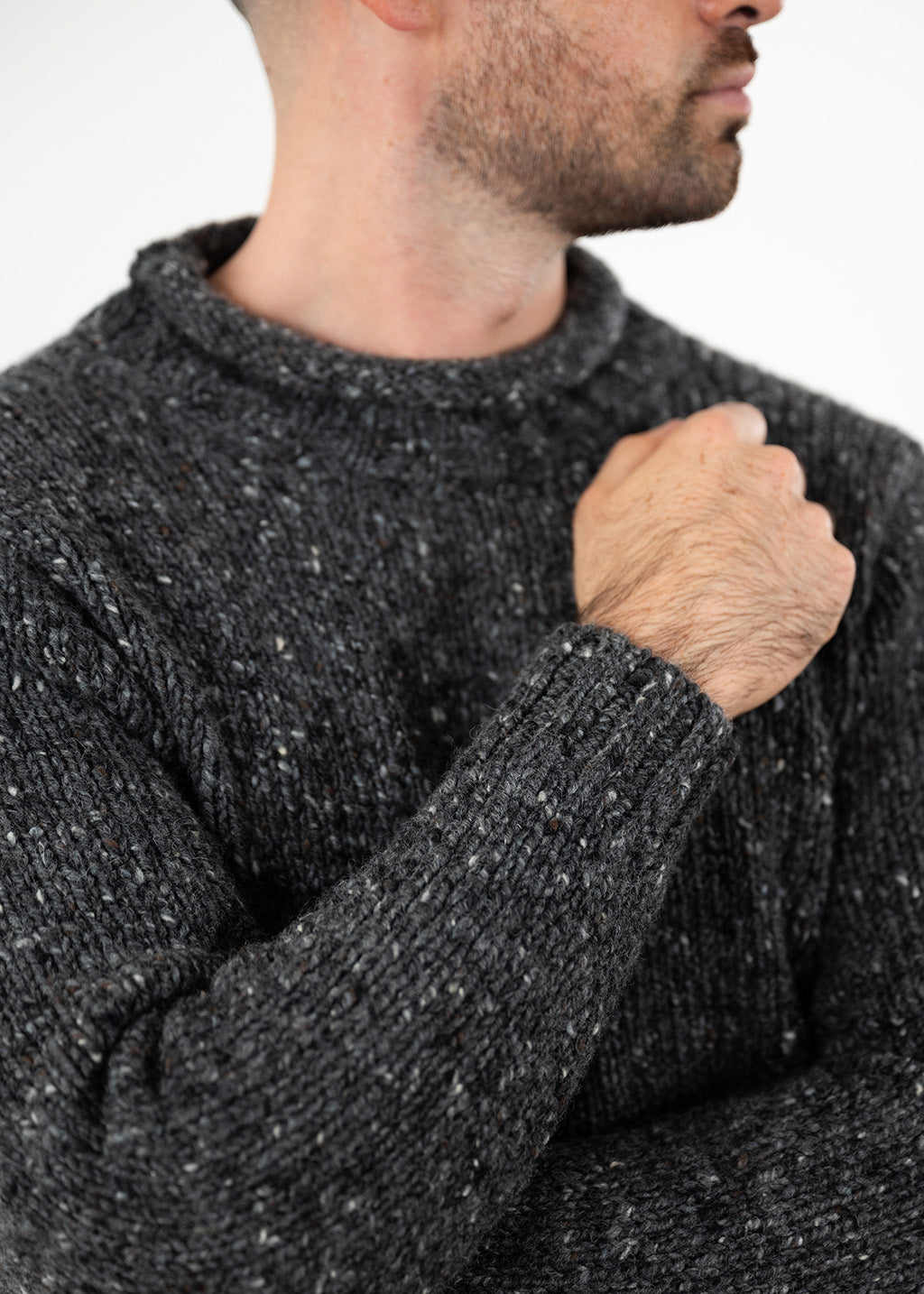 A close-up of a man wearing a dark  gray donegal wool knit sweater, showing the texture of the ribbed rolled edge collar, sleeve, and cuff, with his hand resting on his chest against a plain white background.