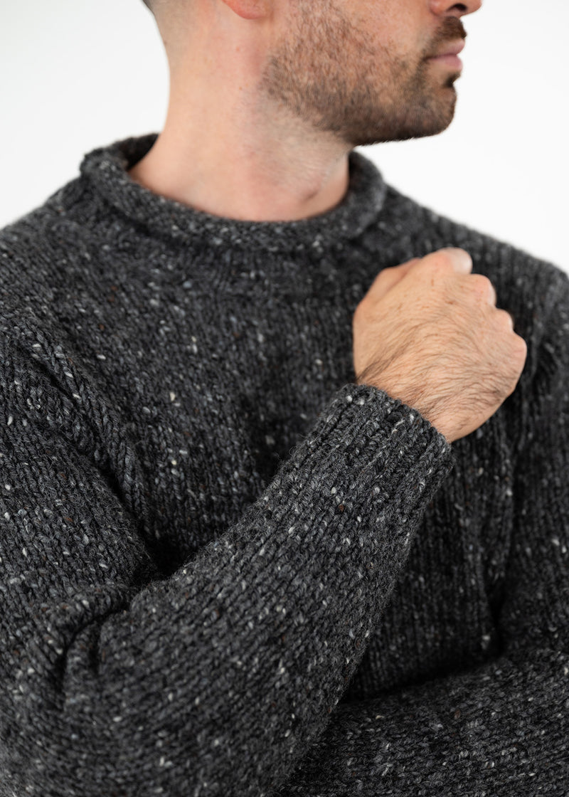 A close-up of a man wearing a dark  gray donegal wool knit sweater, showing the texture of the ribbed rolled edge collar, sleeve, and cuff, with his hand resting on his chest against a plain white background.