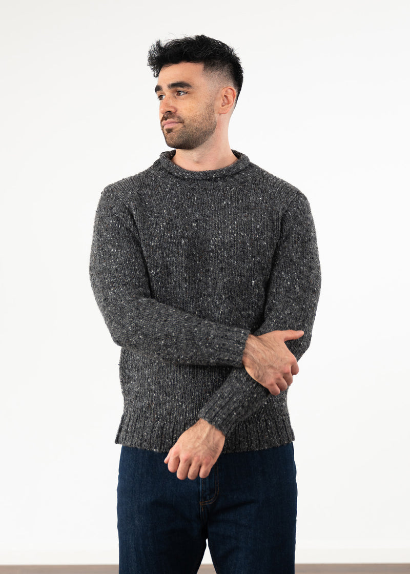 A man wearing a dark gray flecked knit jumper and dark blue jeans, standing against a plain white studio background.