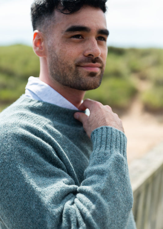 A man wearing a shetland wool sage green knit sweater over a pale blue collared shirt, standing outdoors with a blurred beach dune background.