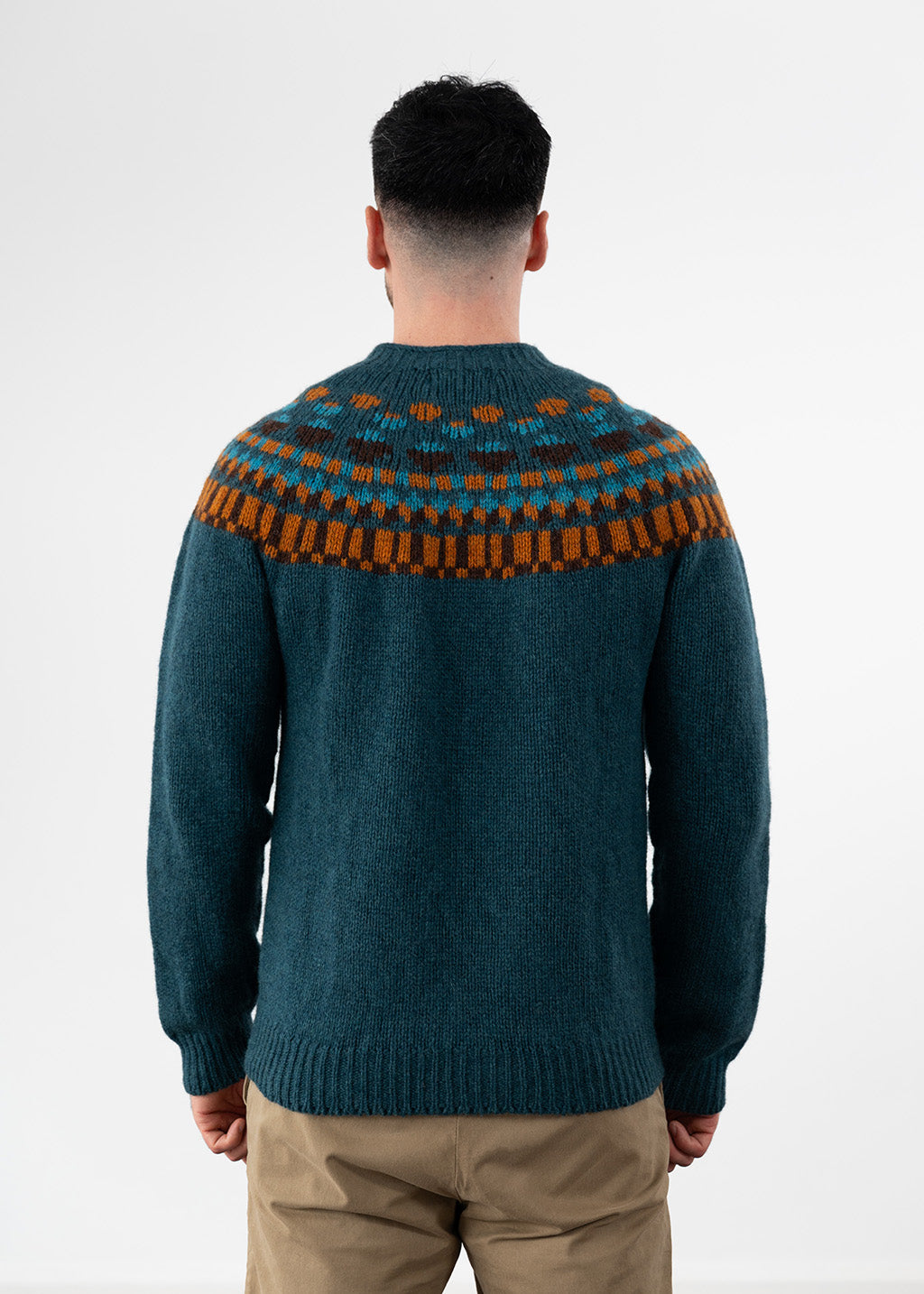 A man viewed from behind wearing a dark teal knit sweater with a brown and orange geometric yoke pattern, paired with khaki pants, against a plain white studio background.