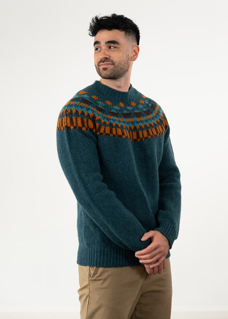 A man wearing a dark teal knit fair isle jumper with a brown and orange geometric pattern across the yoke, paired with khaki trousers, standing against a plain white studio background.