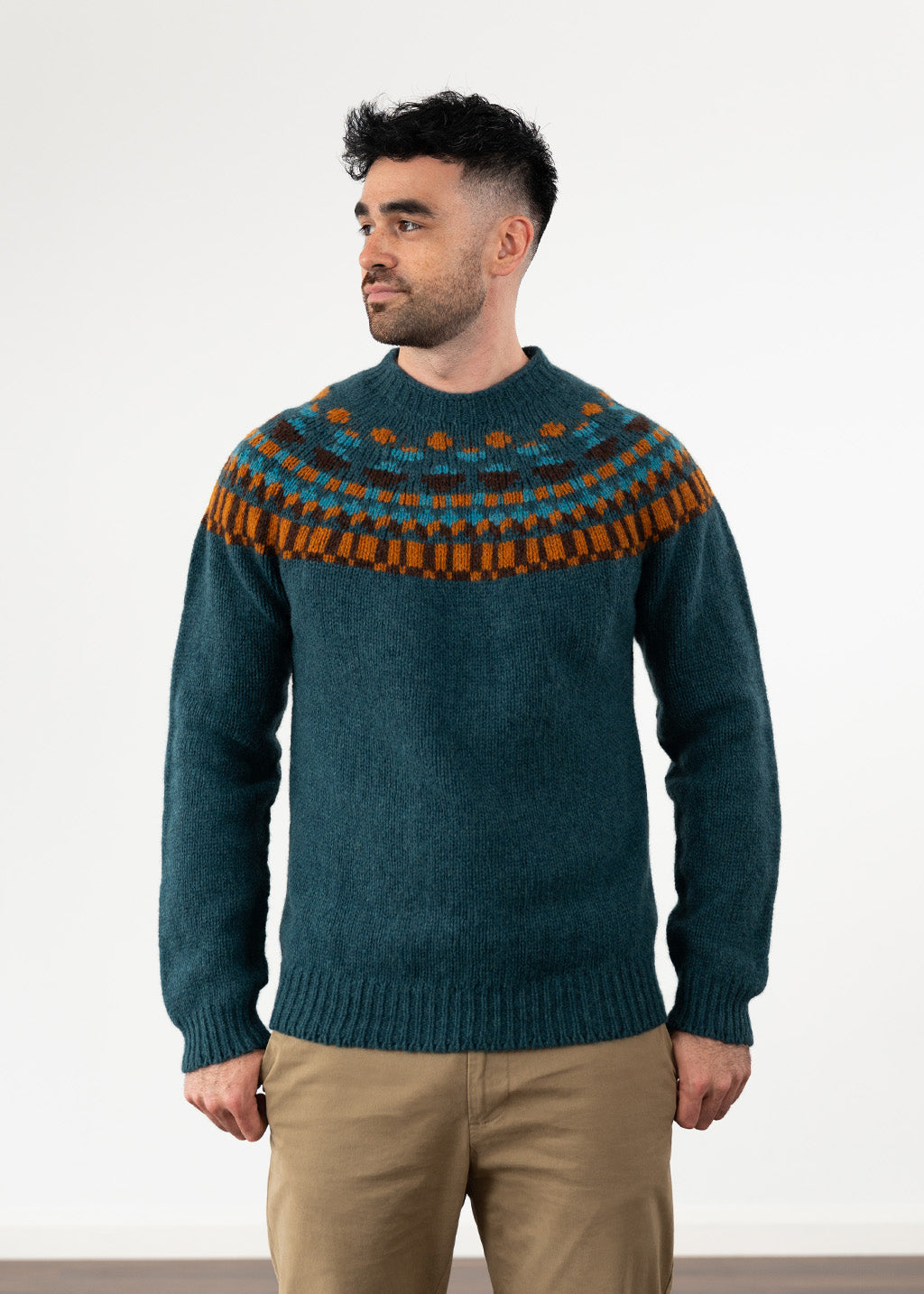 A man wearing a dark teal knit sweater with a brown and orange geometric Fair Isle yoke pattern, paired with khaki chinos, standing against a plain white studio background.