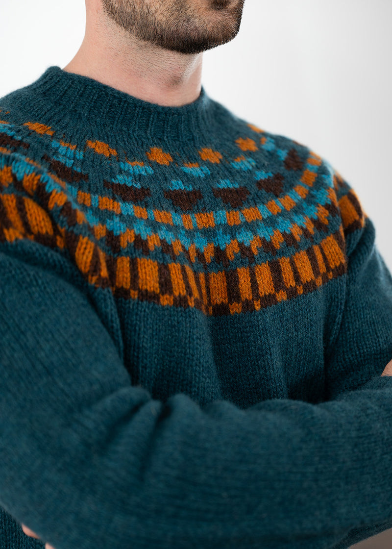 A close-up of a man wearing a dark teal knit sweater with a colourful Fair Isle yoke pattern in orange, brown, and turquoise blue.