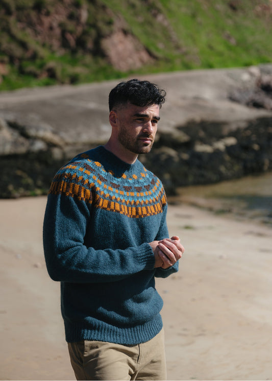A man on a beach wearing a dark teal fair isle knit jumper with orange and brown pattern across the yoke and beige chinos