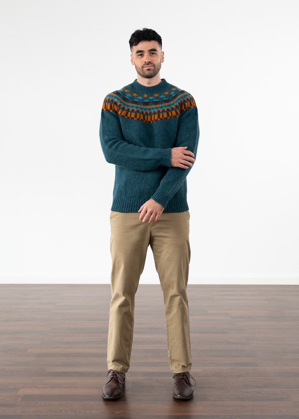 A man wearing a dark teal knit sweater with a multicolour fair isle pattern across the yoke, paired with tan trousers and brown shoes, standing against a white studio background.