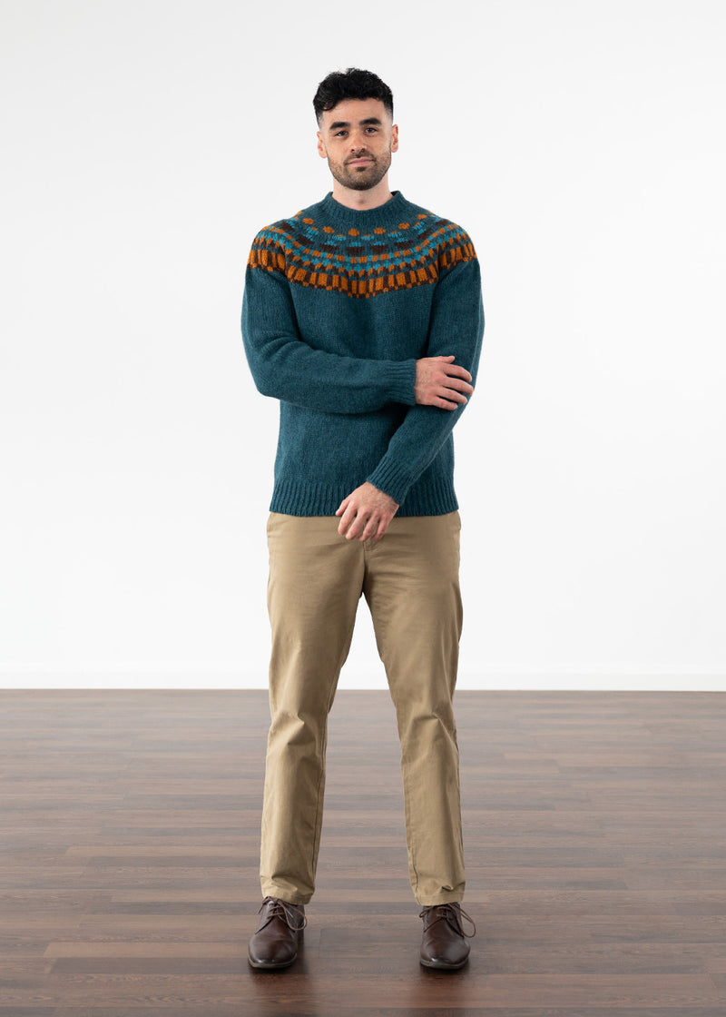 A man wearing a dark teal knit sweater with a multicolour fair isle pattern across the yoke, paired with tan trousers and brown shoes, standing against a white studio background.