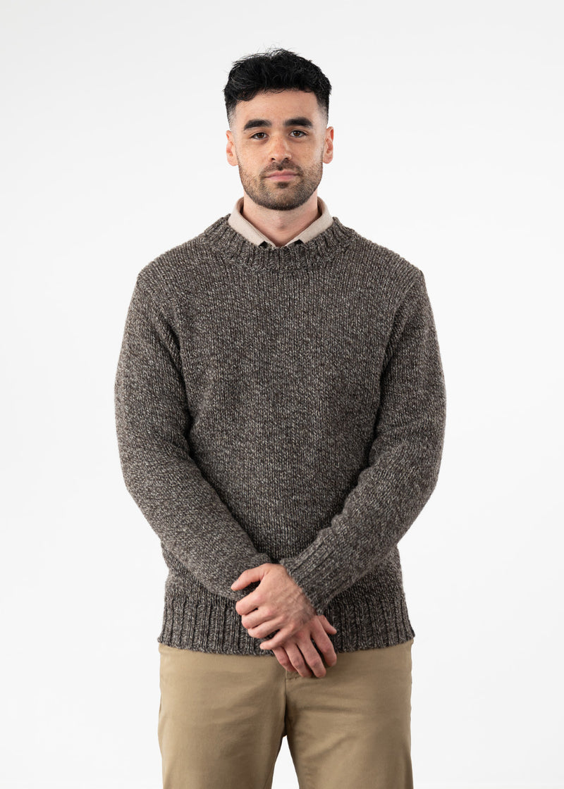 A man wearing a brown marl wool crew neck jumper over a beige shirt with khaki chino trousers, against a white wall.