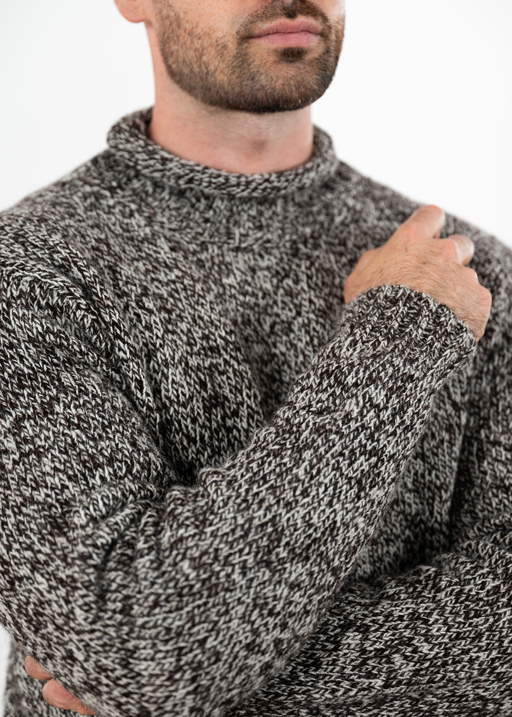 A close-up of a man wearing a thick, textured black and white marled knit jumper, showing the ribbed collar and his hand resting on his chest against a plain white background.
