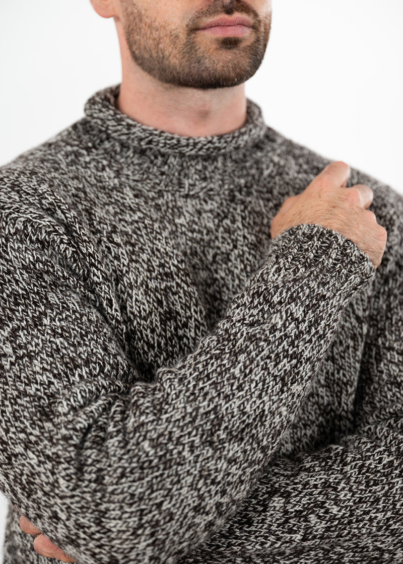 A close-up of a man wearing a thick, textured black and white marled knit jumper, showing the ribbed collar and his hand resting on his chest against a plain white background.