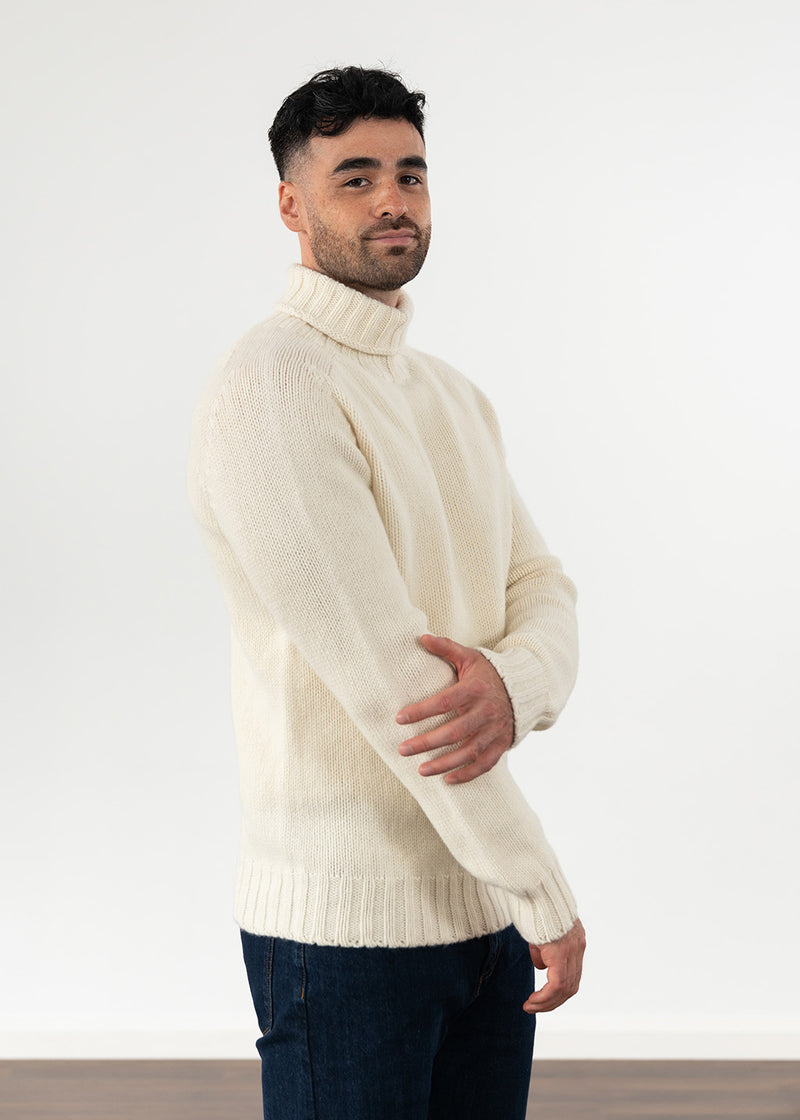 A man wearing a cream-colored, ribbed-knit turtleneck sweater and dark blue jeans, standing against a plain white studio background.