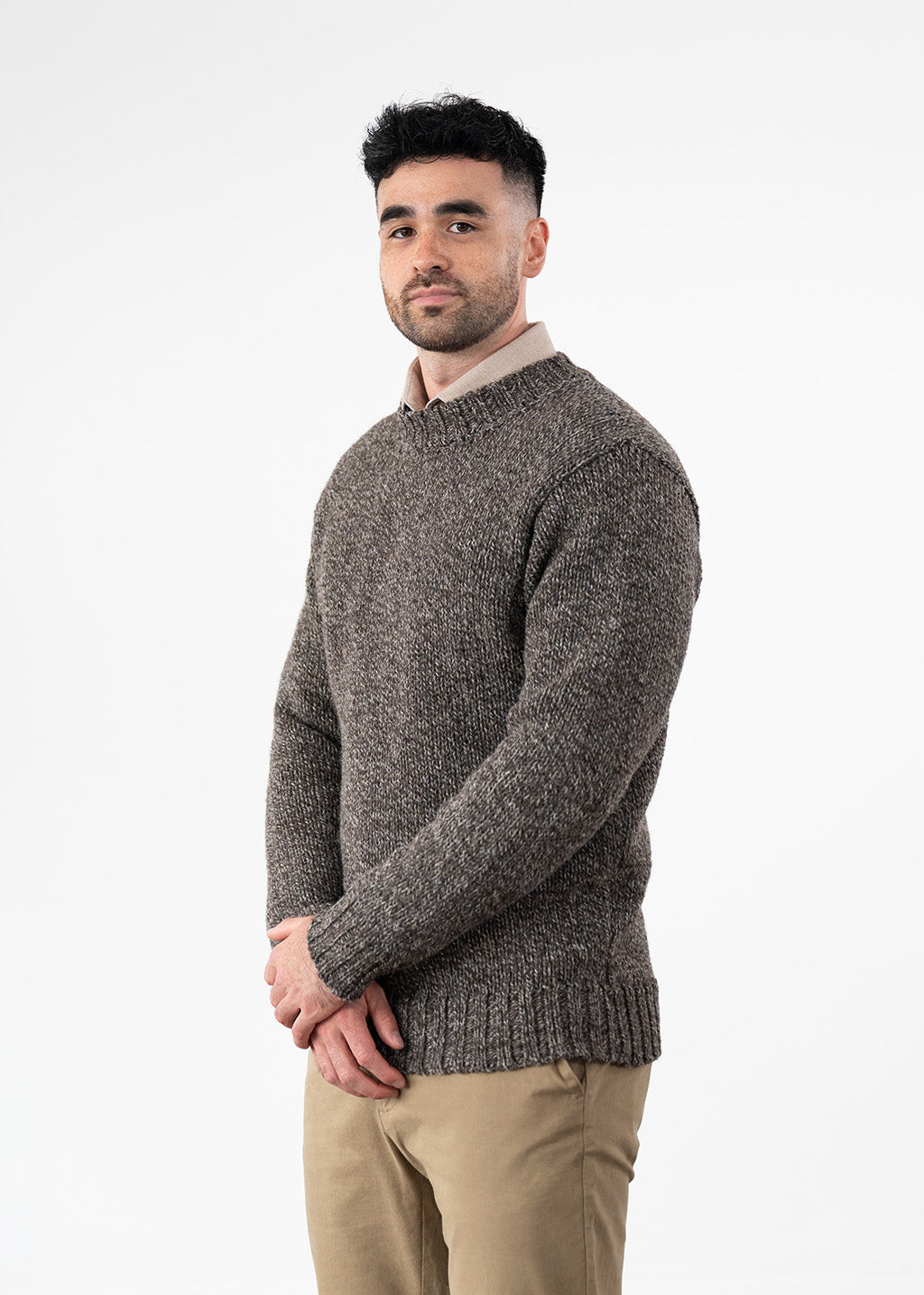 A man wearing a textured, heathered brown knit sweater over a light beige collared shirt and khaki pants, standing against a plain white studio background.