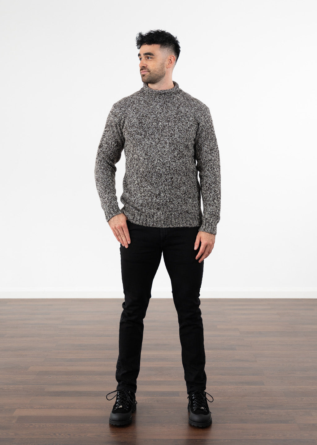 A man wearing a black and ecru marled knit roll neck jumper, black jeans and black boots, standing against a white wall and wooden floor.