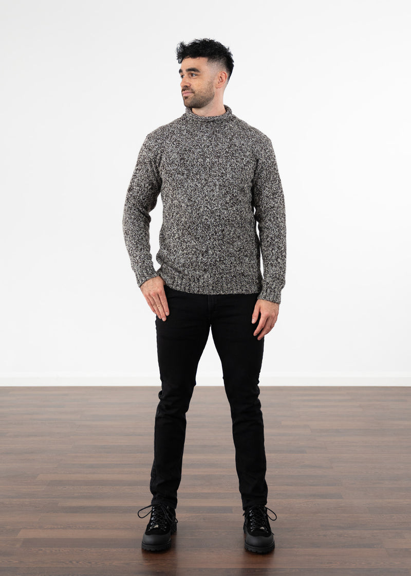 A man wearing a black and ecru marled knit roll neck jumper, black jeans and black boots, standing against a white wall and wooden floor.