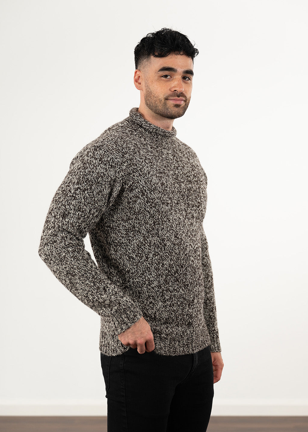 A man wearing a textured, long-sleeve knit sweater in a marled black and undyed ecru pattern, paired with black jeans, standing against a plain white wall.