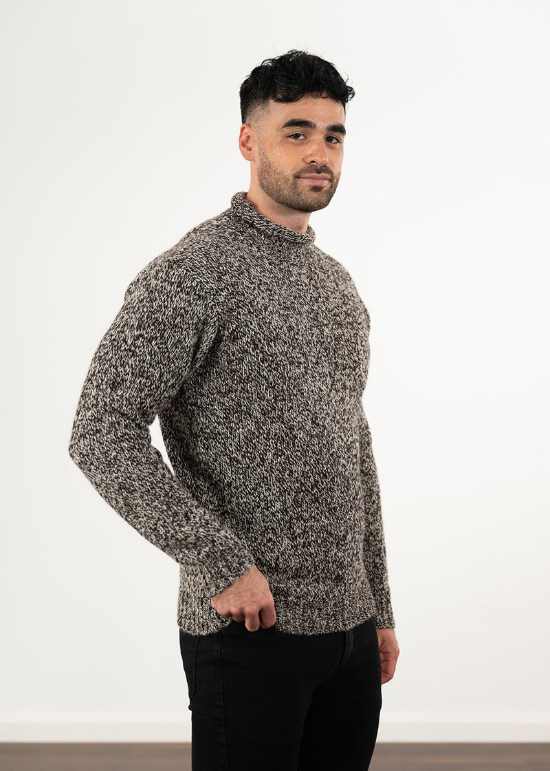A man wearing a textured, long-sleeve knit sweater in a marled black and undyed ecru pattern, paired with black jeans, standing against a plain white wall.