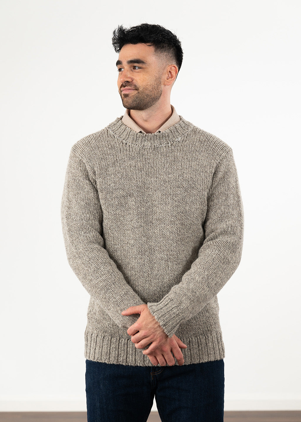 A man wearing a light grey-brown melange knit sweater over a beige collared shirt and dark blue jeans, standing against a plain white wall.