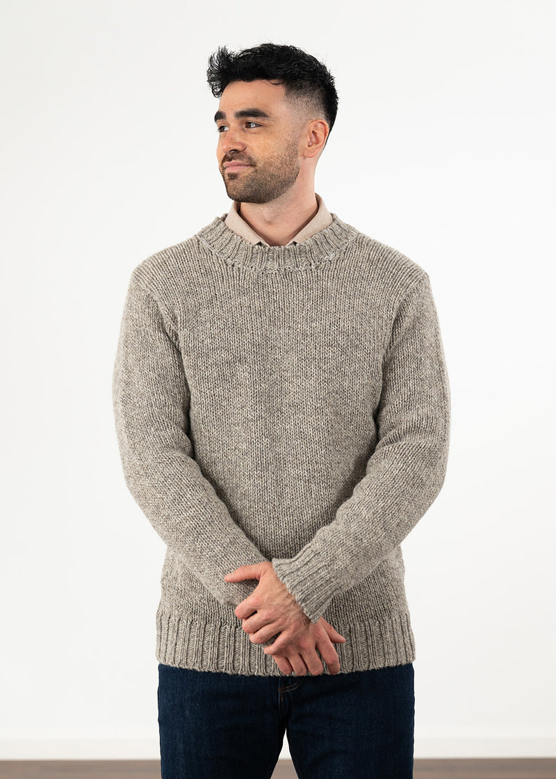 A man wearing a light grey-brown melange knit sweater over a beige collared shirt and dark blue jeans, standing against a plain white wall.