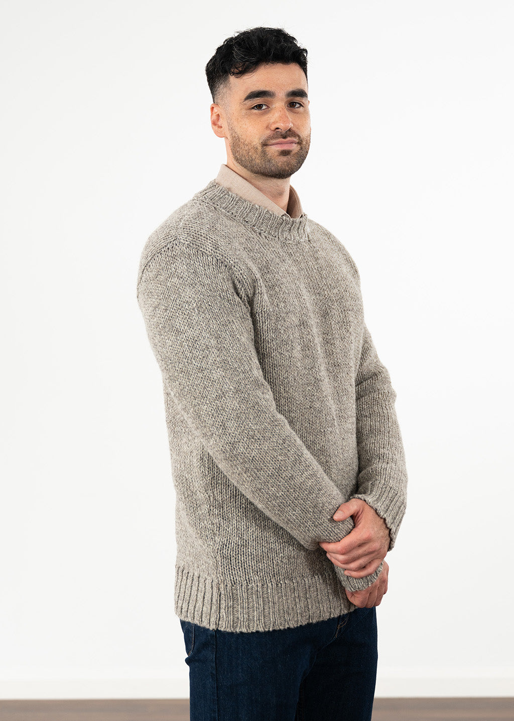 A man wearing a light gray-brown textured knit crewneck sweater, paired with a beige collared shirt and dark blue jeans, standing against a plain white wall.