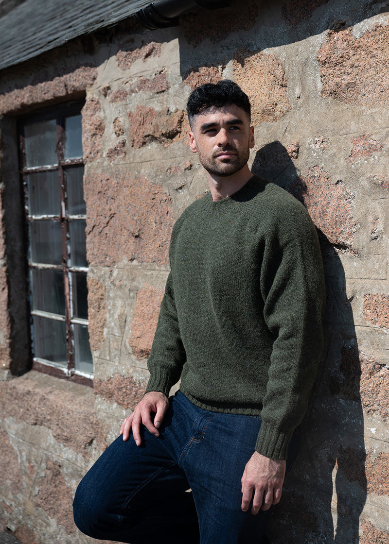 A man wearing an olive green knit jumper and dark blue jeans leans against a rustic stone cottage wall outdoors.