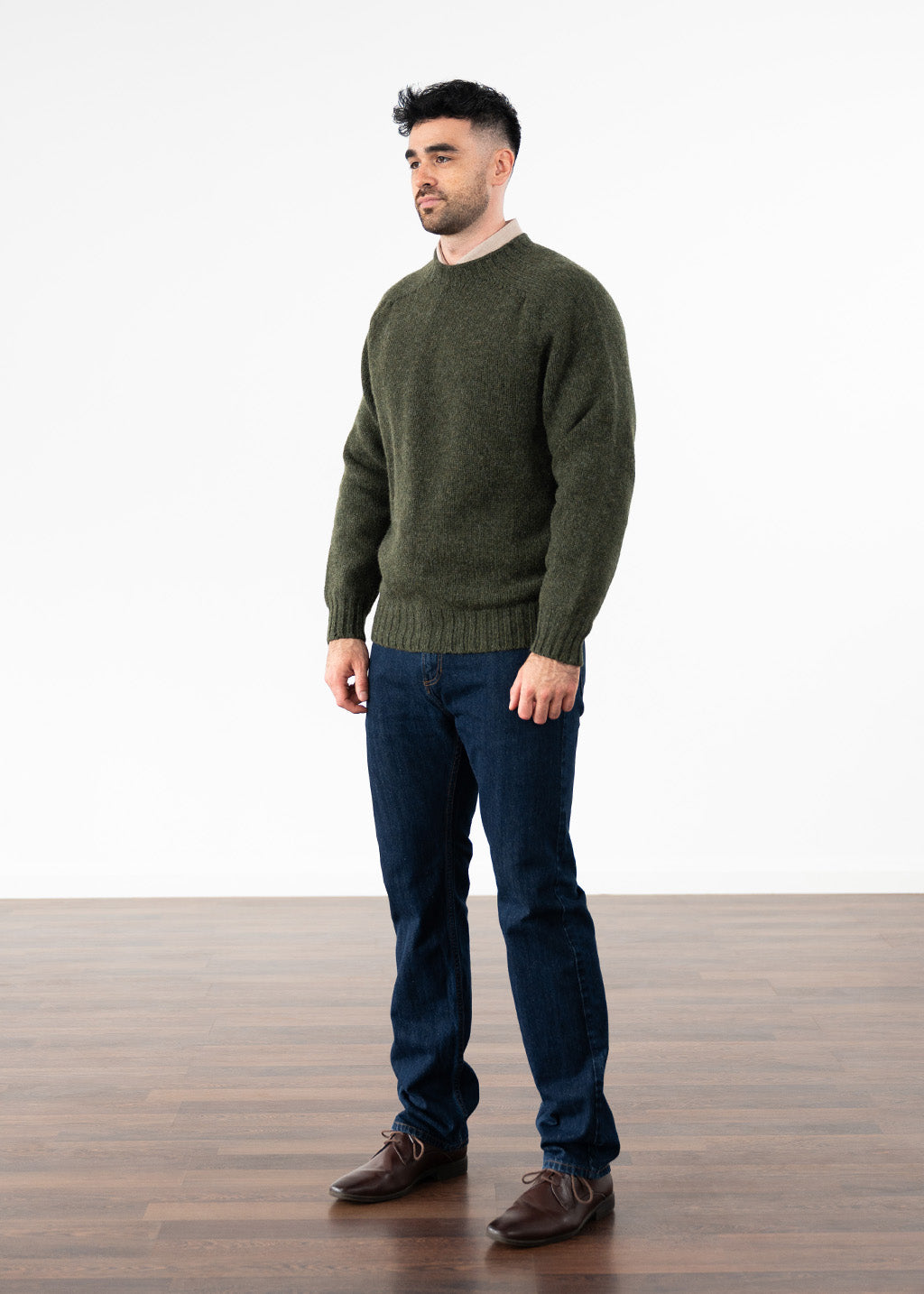 A man wearing a country green knit crew neck jumper, dark blue jeans, and brown leather shoes, standing against a white studio background with a wooden floor.