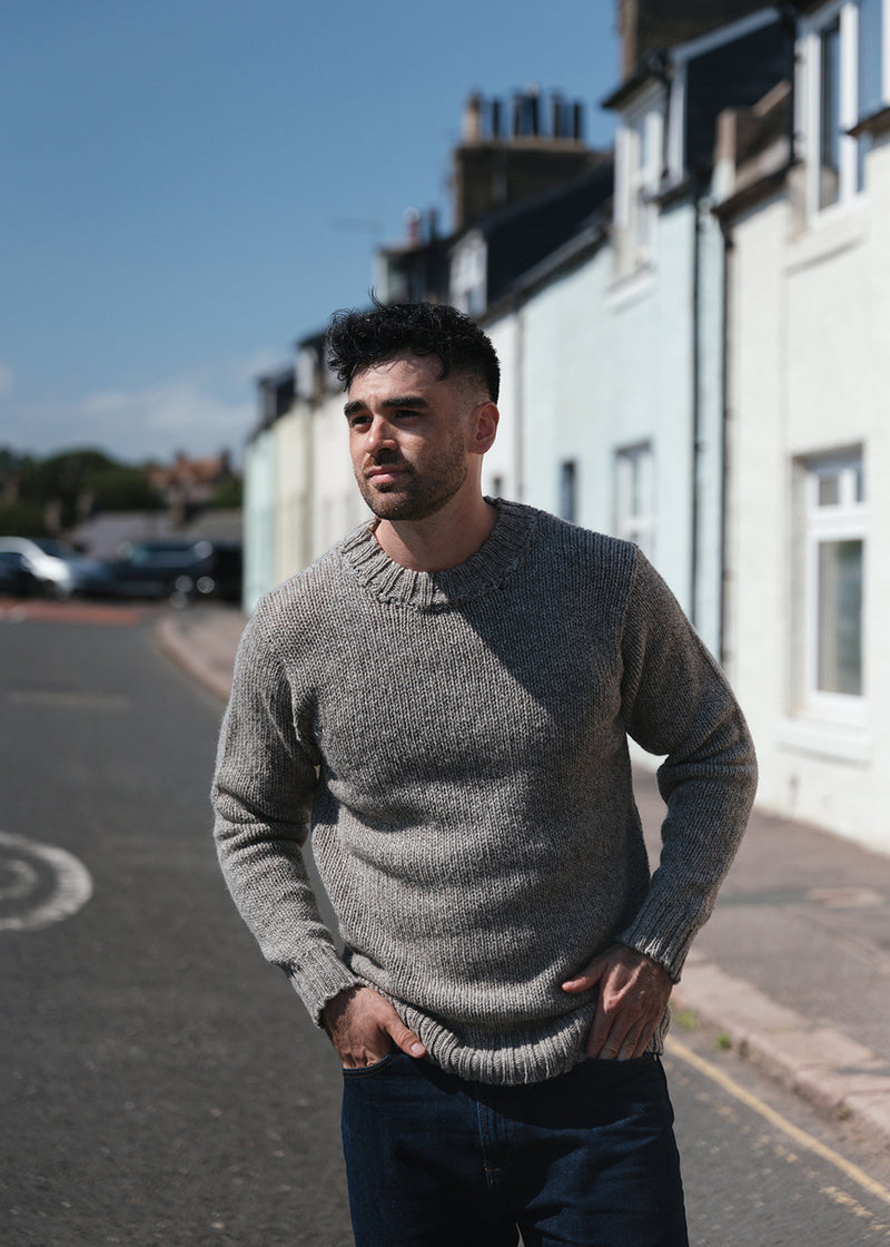 A man wearing a grey-brown knitted crew neck jumper and dark denim jeans, standing on a sunny street with pastel coloured cottages in the background.