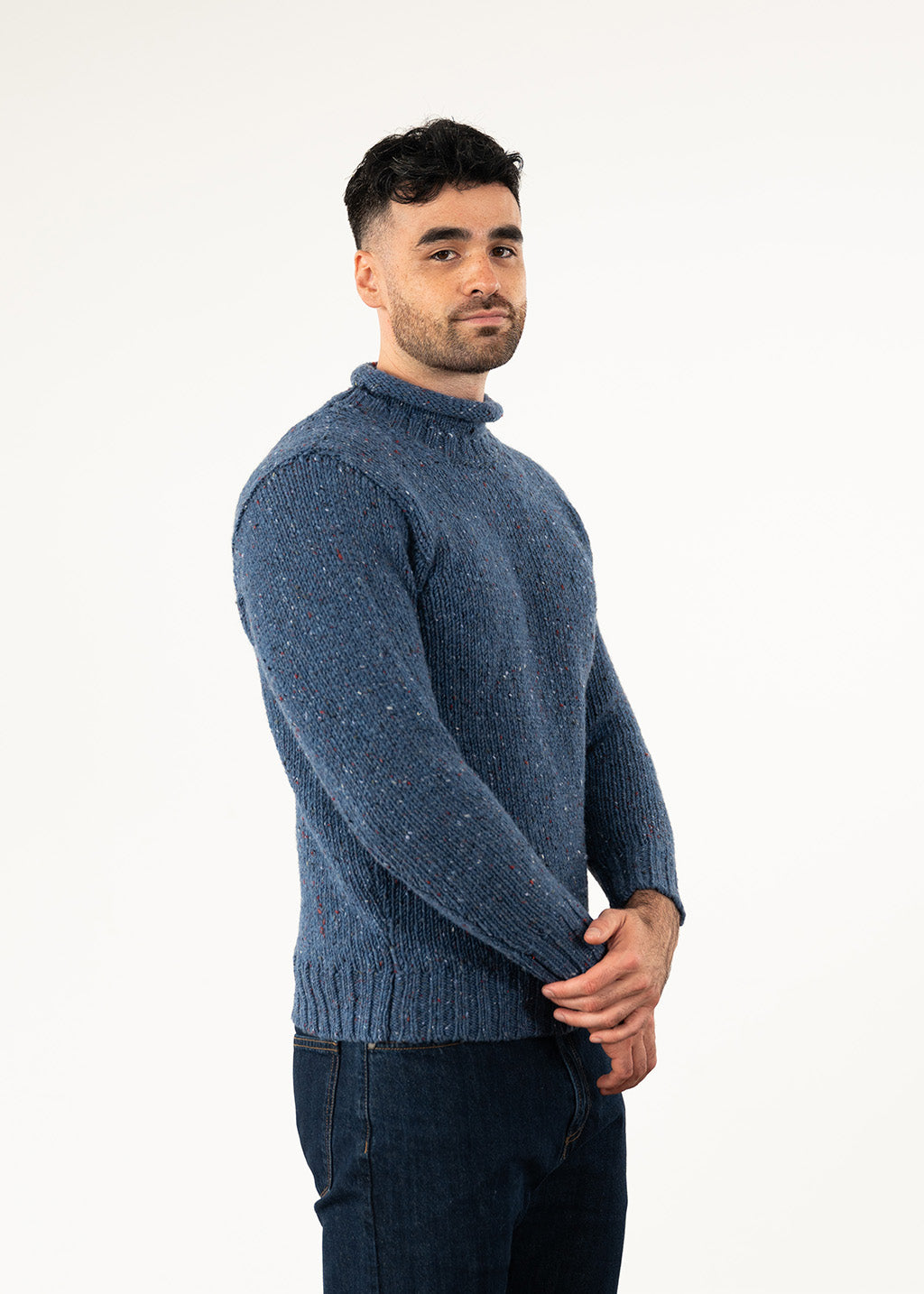 A man wearing a textured blue roll neck jumper and blue jeans, standing against a plain white studio background.