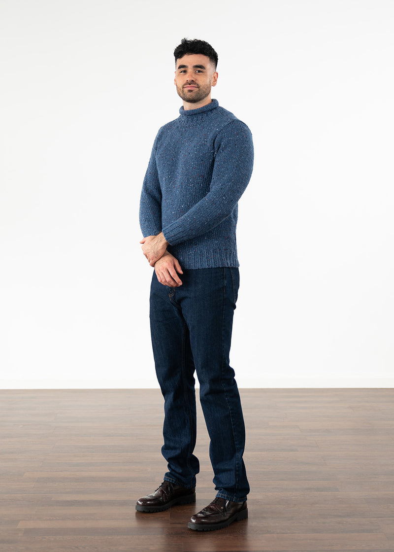 A man wearing a blue knit jumper, dark blue jeans, and dark burgundy leather shoes, standing against a white studio background with a wooden floor.