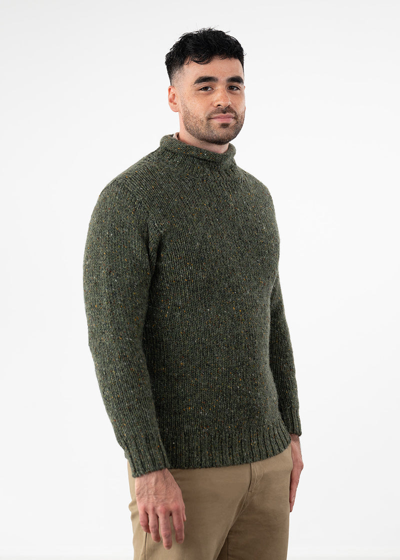 A man wearing a dark olive green textured knit turtleneck sweater and khaki trousers, standing against a plain white studio background.