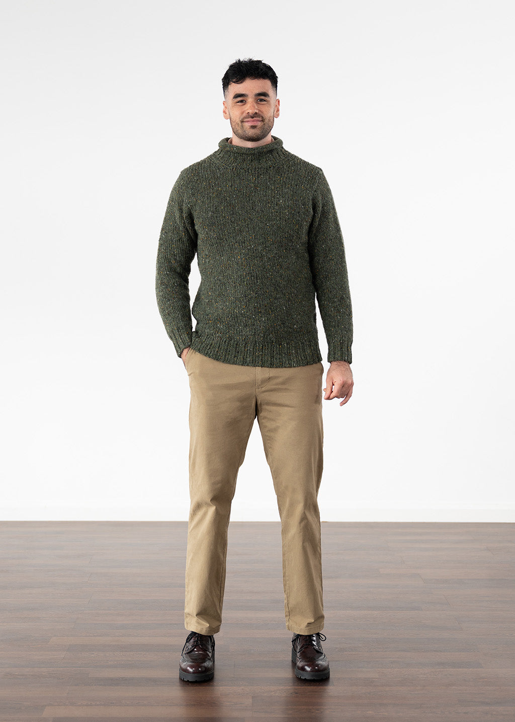 A man wearing a dark olive green knit turtleneck jumper, beige trousers, and dark brown leather shoes, standing against a white studio background with a wooden floor.