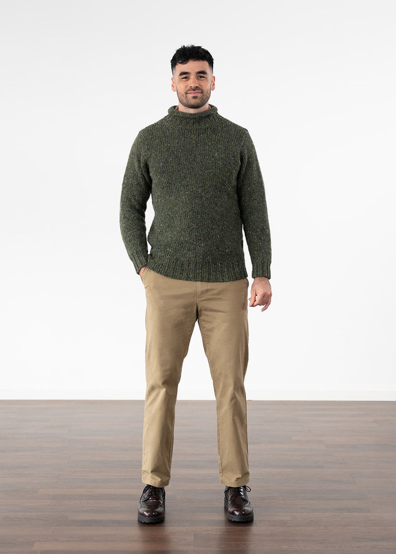 A man wearing a dark olive green knit turtleneck jumper, beige trousers, and dark brown leather shoes, standing against a white studio background with a wooden floor.