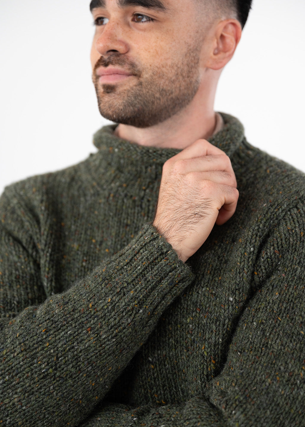 A close-up of a man wearing a dark olive green wool turtleneck sweater with subtle multicolour flecks, set against a plain white background.